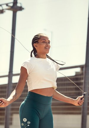 White cropped t-shirt and green high-waisted leggings with floral pattern. Jump rope held in hands; braided hair tied back in a ponytail.