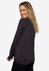 Langärmlige Bluse aus schwarzem Stoff mit einem sich wiederholenden Muster aus rosa und weißen Buchstaben. Lockerer Schnitt mit abgerundetem Saum und weicher Textur.