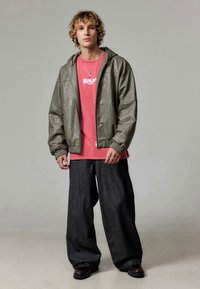 Joven con cabello rizado lleva una chaqueta verde texturizada oversized, una camiseta rosa con la palabra "Berlín", pantalones anchos negros, anillos y zapatos oscuros, de pie.