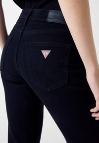 Jeans in denim nero con una toppa triangolare rossa e bianca sul taschino posteriore e una cintura nera opaca.