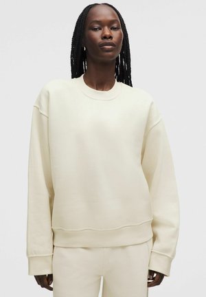 Kobieta z długimi warkoczami w kremowej, oversize'owej bluzie i pasujących spodniach, stojąca na jednolitym jasnym tle.