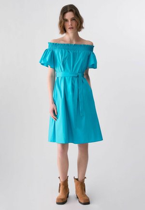 Kobieta w turkusowej sukience za kolano odkrywającej ramiona, z paskiem w talii i brązowych botkach, stojąca na jednolitym tle.