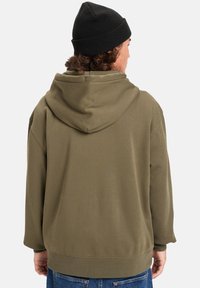 Sudadera verde oliva con una gran capucha suave. Corte holgado, mangas largas, puños y dobladillo de canalé, textura suave, usada sobre un pantalón vaquero azul.