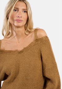 Maglione marrone a maglia con design off-shoulder, bordi sfrangiati e finitura testurizzata. Materiale morbido con una vestibilità ampia e rilassata.