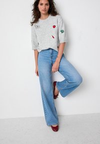 Maglione grigio chiaro lavorato a maglia con frutta ricamata, abbinato a jeans di denim blu larghi con orlo sfilacciato e scarpe rosse basse.