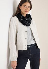 Veste courte gris clair avec fermeture à bouton, tissu doux ; écharpe noire avec motif blanc ; portée sur un haut gris et un jean sombre.