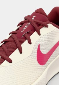 Nike-sko med en hvit teksturert overdel, maroon aksenter, rosa logo og maroon lisser, med et lett design og pustende materiale.