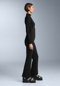 Černý, těsně přiléhající overal s rolákem, který má nastavitelné popruhy a viditelné švy, doplněný o černé lyžařské boty a kovové spony.