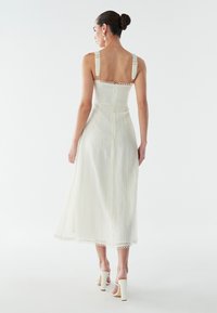 Balta midi kleita ar fakturētu audumu, mežģīņu lencītēm un apaļiem akcentiem. Raksturīga pieguļošs korsete un izplests svārku plats ar viļņotu apmali.