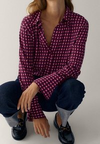 Femme accroupie portant une chemise à motifs géométriques rouge et blanc, un jean foncé à revers et des mocassins noirs avec des accents dorés.