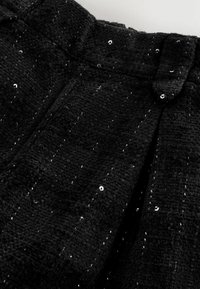 Tissu noir texturé avec une broderie verticale subtile et de petites paillettes noires brillantes dispersées, comportant un détail de passant de ceinture.