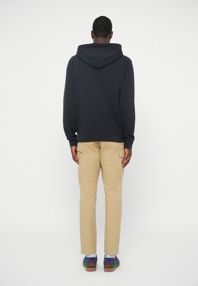 Ναυτική φούτερ με μεγάλη κουκούλα, συνδυασμένο με μπεζ cargo παντελόνι. Τα παπούτσια διαθέτουν πράσινες λεπτομέρειες και πολυχρωματικά στοιχεία. Απλό φόντο.