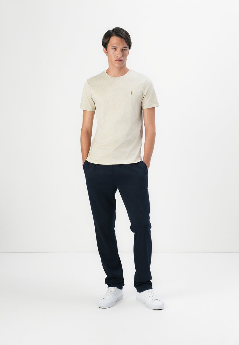 Camiseta de cuello redondo color beige con un pequeño logo, combinada con pantalones jogger azul marino y zapatillas blancas. Un look sencillo y casual con un ajuste relajado.