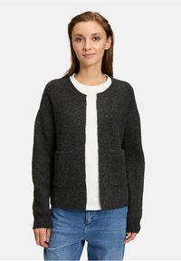 Ciemnoszary kardigan wykonany z teksturowanej wełny, z długimi rękawami, dwoma przednimi kieszeniami i okrągłym dekoltem. Nosi się go na białej bluzce.