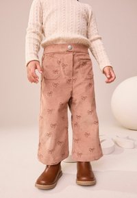 Pantaloni in corduroy rosa con delicati motivi a fiocchi, gamba ampia, due tasche frontali e chiusura a scatto anteriore. Abbinati a stivali alla caviglia marroni.