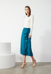 Gonna midi in satin teal con dettaglio arricciato in vita, abbinata a una blusa crema a maniche lunghe e tacchi dorati con cinturini. Tessuto liscio.