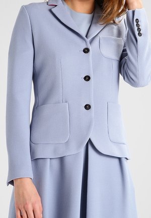 Frau trägt einen hellblauen strukturierten Blazer mit drei schwarzen Knöpfen und passendem Rock, Hand nahe am Gesicht, vor einfarbigem Hintergrund.