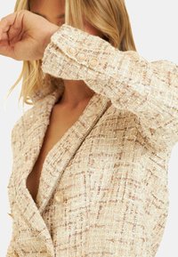 Blazer en tweed beige clair avec un tissage texturé, des revers en notch et des accents de boutons dorés sur les poignets.