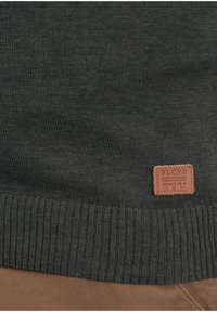 Pull en maille gris foncé avec un ourlet côtelé et une étiquette de marque en cuir marron. Texture lisse avec un design simple et classique.