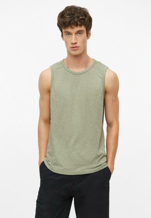 Junger Mann mit lockigem Haar, der ein hellgrünes ärmelloses Shirt und eine schwarze Hose trägt, steht mit den Händen in den Taschen vor einem weißen Hintergrund.