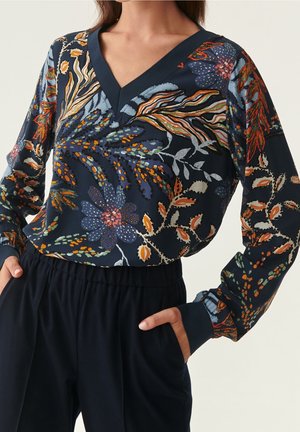 Vrouw met een marineblauwe blouse met V-hals en lange mouwen, bedrukt met kleurrijke abstracte bloemenpatronen, gecombineerd met zwarte, hooggetailleerde broek.