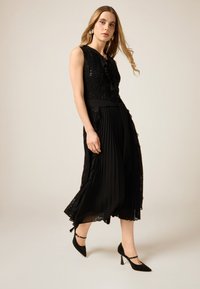 Czarna sukienka z cekinowym topem ozdobionym falbanami i plisowaną spódnicą, zestawiona z czarnymi szpiczastymi szpilkami i paskowymi akcentami.