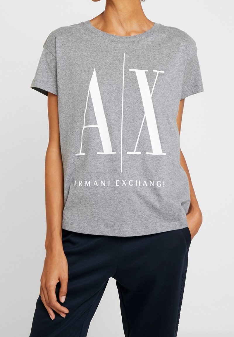 Femme portant un T-shirt gris à manches courtes avec un grand logo blanc "A | X" et le texte "Armani Exchange", associé à un pantalon foncé.
