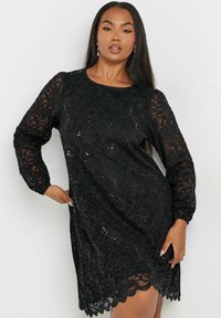 Musta pitsimekko pitkillä hihoilla, napsauttamalla reunoja ja ripoteltuja niittejä. Kankaassa on kukkakuvio, joka lisää tekstuuria ja yksityiskohtia.