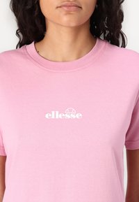 Lyserød bomulds t-shirt med rund halsudskæring, prydet med et hvidt "ellesse" logo centralt på brystet. Korte ærmer, glat tekstur.