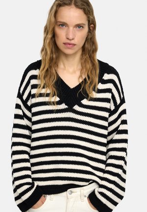 Młoda kobieta o długich, falowanych blond włosach, ubrana w czarno-biały sweter w paski i białe spodnie, z rękami w kieszeniach, na neutralnym tle.