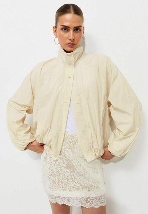 Donna che indossa una giacca oversize beige chiaro, top bianco e una minigonna bianca in pizzo e paillettes, in piedi con le mani sui fianchi davanti a uno sfondo bianco.