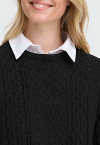 Maglione nero lavorato a maglia con un motivo a trecce testurizzato, collo rotondo e una camicia bianca con colletto visibile sotto.