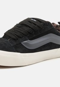 Fekete suede sneaker, szürke bőr kiegészítővel, kerek fűzőkkel, párnázott nyakkal és texturált gumitalppal; finom perforálásokkal a légáteresztésért.