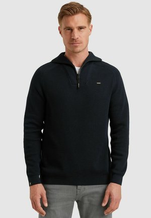Mann trägt schwarzen, gerippten Halb-Reißverschluss-Pullover und graue Hose, steht vor einfarbig hellem Hintergrund.