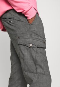 Šedé cargo kalhoty s kostkovaným vzorem, obsahující boční kapsu se zmáčknutím a malou štítkovou nášivku, nošeny pod růžovou košilí s dlouhým rukávem.