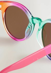 Lunettes de soleil colorées avec monture transparente verte et rose, verres marron et une forme ronde unique. Dotées d'une branche rose flexible.