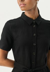 Camicia nera con bottoni in tessuto leggero, dotata di maniche corte, colletto e dettaglio in vita annodato per un look aderente.