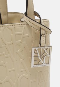 Beżowa torba typu tote z wytłoczonym wzorem logo, gładką fakturą i krótkimi czarnymi uchwytami. Zawiera srebrną zawieszkę z logo przymocowaną do czarnego paska.