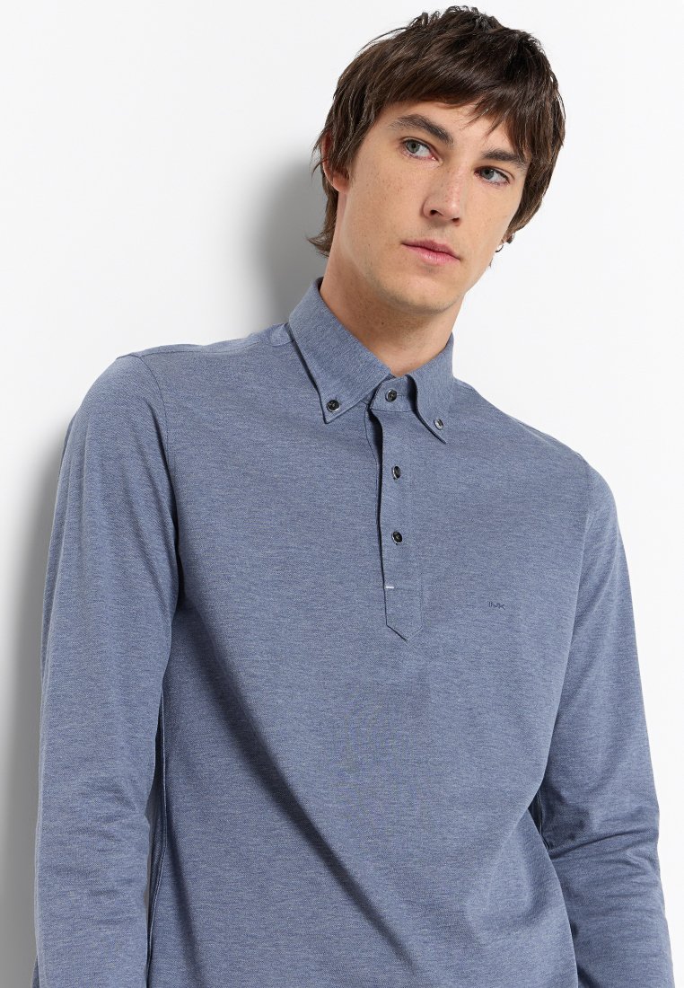 Joven con cabello corto castaño, vistiendo una camisa de polo azul con mangas largas y botones, mirando ligeramente hacia su derecha contra un fondo blanco.