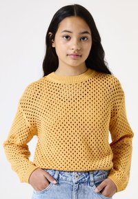 Pull en tricot jaune à motif en maille ouverte et col rond. Manches longues et tissu texturé. Porté avec un jean en denim clair.