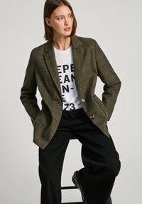 Blazer a quadri verdi con rever, chiusura a un bottone, indossato sopra una t-shirt bianca con grafica e pantaloni larghi neri. Tessuto con una texture liscia.