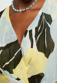 Gros plan d'une personne à la peau foncée portant une robe fleurie avec des motifs jaunes, noirs et bleu clair, ainsi qu'un collier de perles bleu.