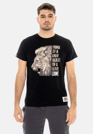 Jonge man draagt een zwart t-shirt met leeuwafbeelding en tekst "Mind of a lion heart of a king Leone 1947" staand tegen een witte achtergrond.