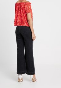 Kvinde står, iført rød off-shoulder blomstret top, højhalsede sorte vide bukser og hvide højhælede sandaler, med ryggen til.