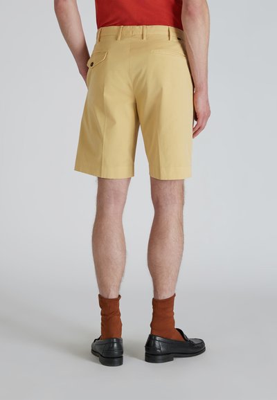 Pantalones cortos de algodón amarillos con corte recto, bolsillo trasero y trabillas, combinados con calcetines marrones y mocasines negros, vistos desde atrás.