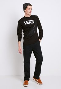 Sort langærmet skjorte med hvid "VANS" logo, sort bumbags, sorte bukser og orange sneakers med hvide detaljer, sat mod en ensfarvet baggrund.