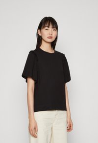 Svart t-skjorte med korte ermer, med volanger på skuldrene, rund halsutskjæring og rett hem. Stoffet ser mykt og glatt ut.