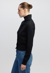 Schwarzer Strickpullover mit hohem Kragen, der subtile silberne Sprenkel aufweist. Das Design beinhaltet fransige Säume und lange Ärmel. Kombiniert mit blauen Jeans.