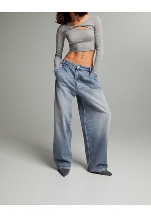 Femme portant un crop top gris à manches longues et un jean bleu délavé, taille haute et ample, les mains dans les poches, debout sur un fond gris.