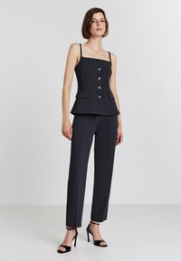 Donna che indossa un top nero senza maniche con bottoni e pantaloni affusolati coordinati, con sandali neri con tacchi alti, in piedi contro uno sfondo semplice.